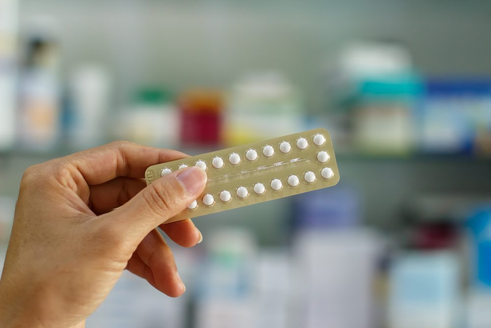 A hand holding a strip of contraceptive pills in front of pharmacy shelves