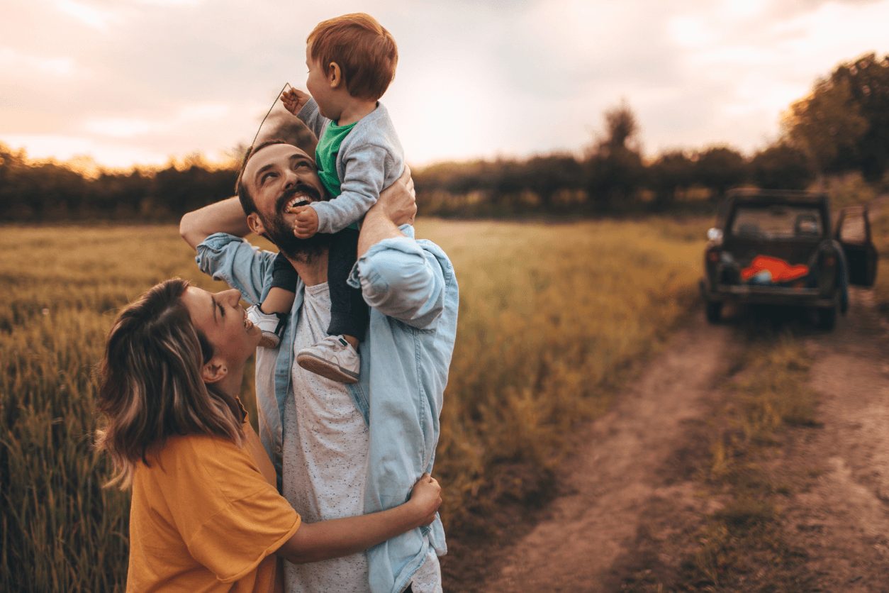 A man carrying a small child on his shoulders, hugged by a woman, in a field at sunset